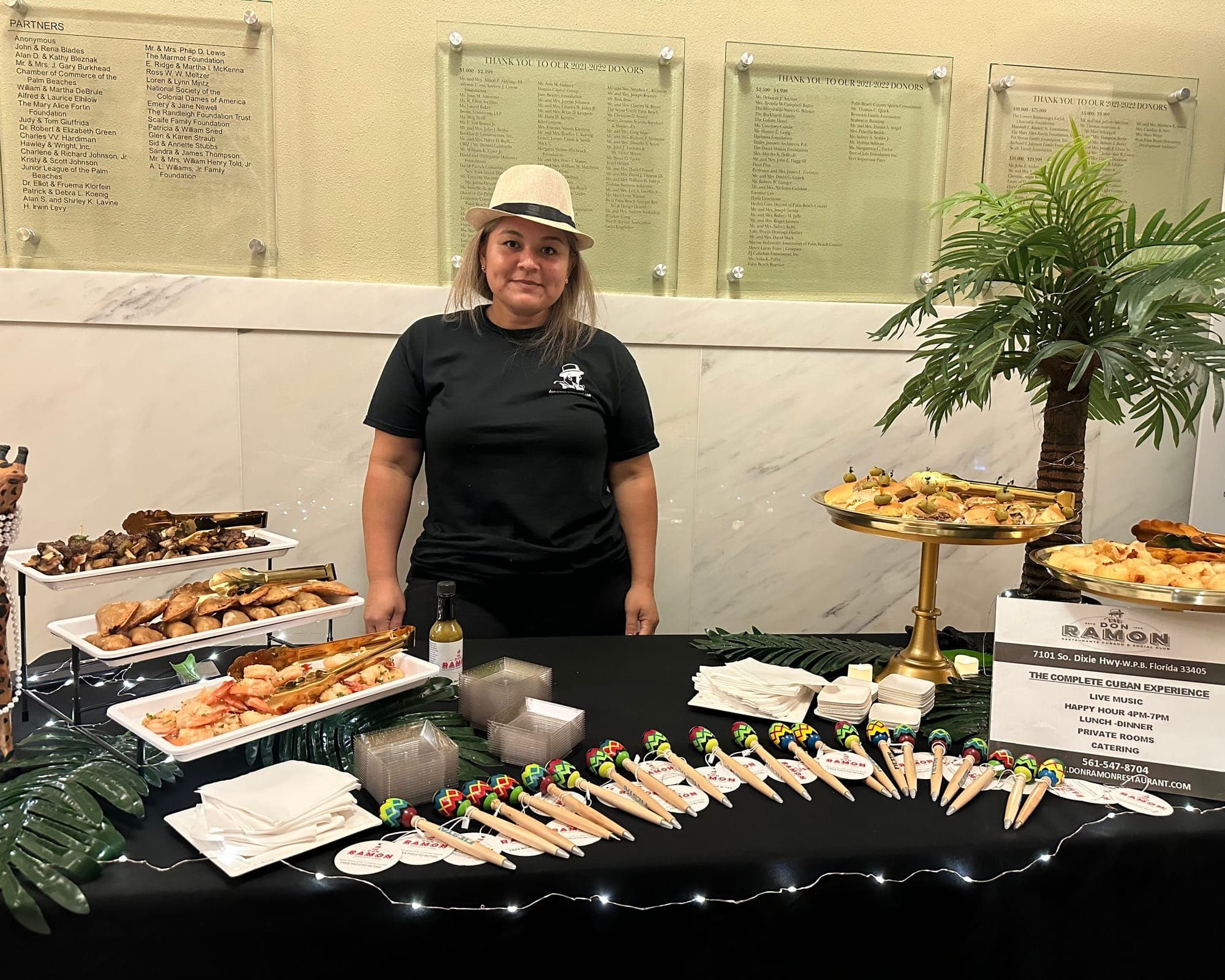 A Don Ramon server in a straw fedora behind a buffet of empanadas, pastelitos and skewers on a black table strung with lights