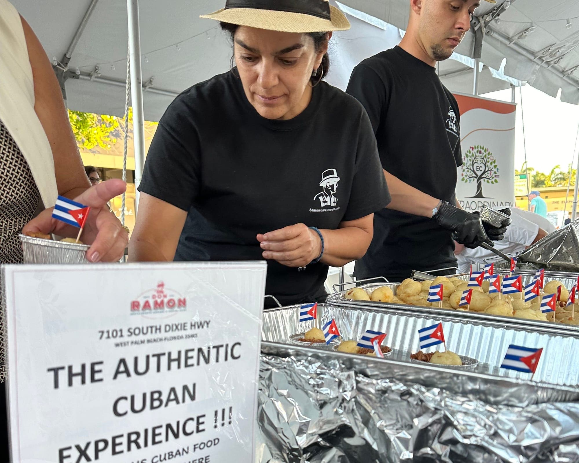 A Don Ramon server in a straw fedora plating croquetas from foil trays topped with little Cuban flags, beside a sign reading The Authentic Cuban Experience