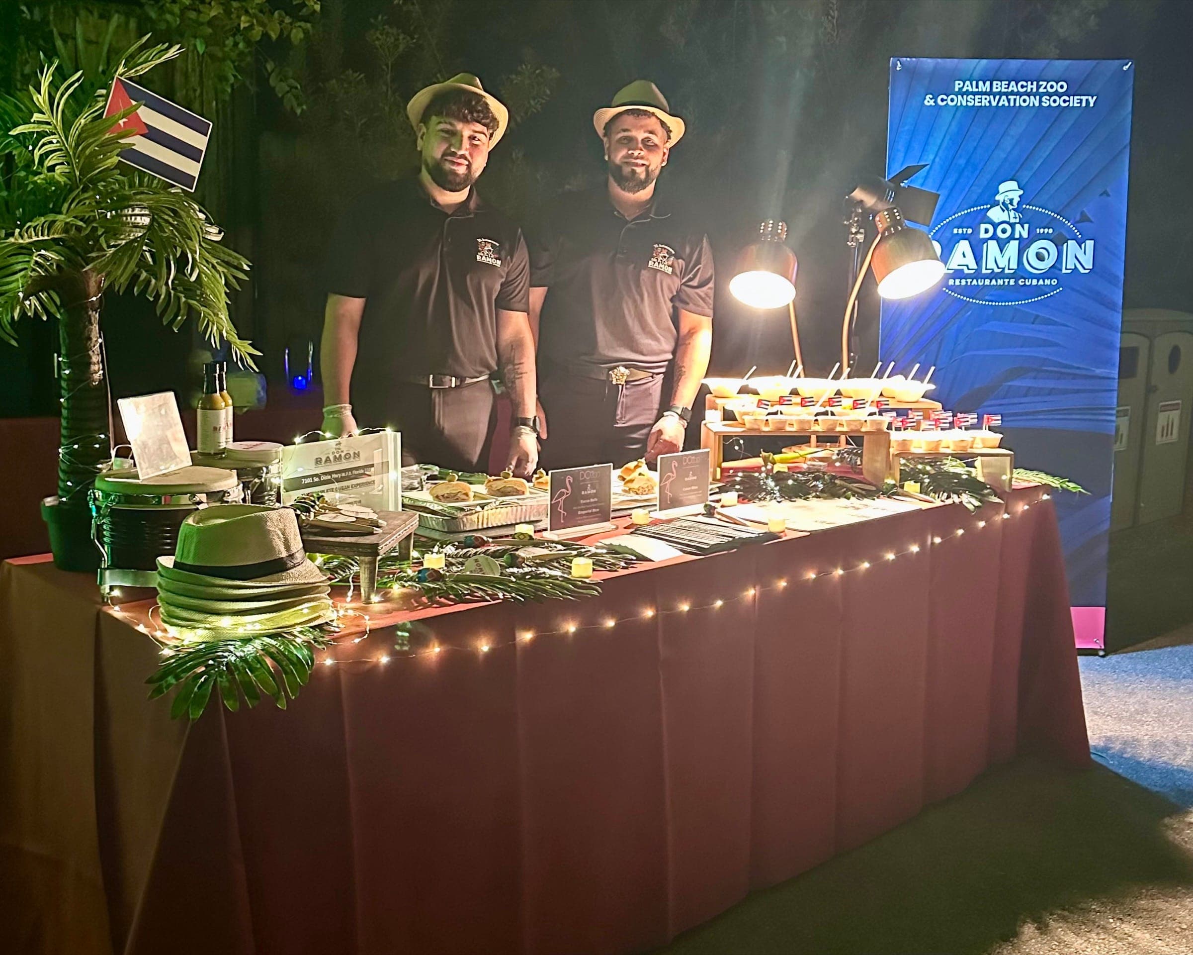 Two Don Ramon servers in fedoras behind a string-lit catering table at a Palm Beach Zoo and Conservation Society evening event