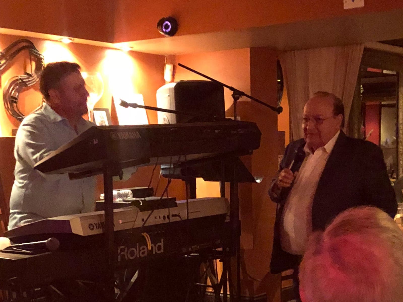 A keyboardist plays a Roland and Yamaha stack while a singer in a dark blazer sings into a microphone for the dining room