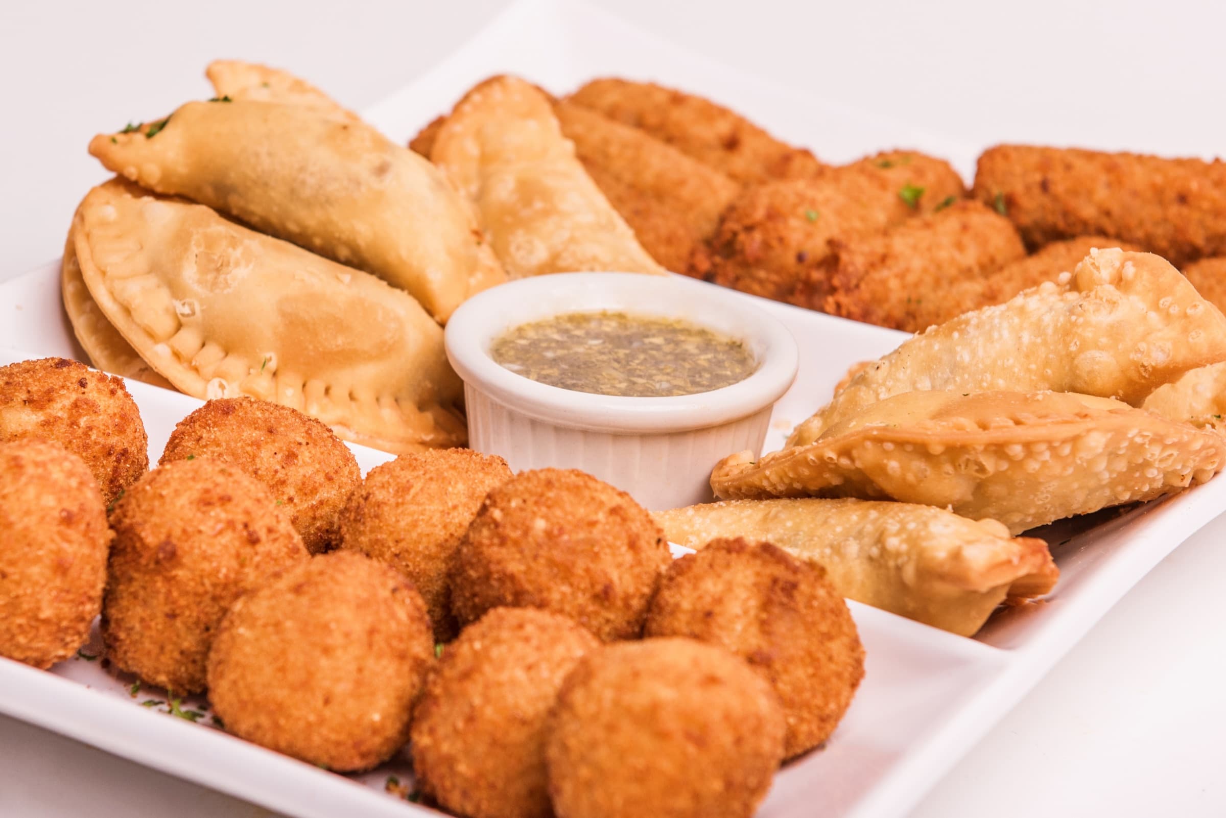 An appetizer platter of croquetas, empanadas and fried dumplings around a cup of mojo